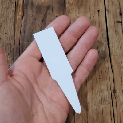 A hand holds a white, flat Plastic Plant Marker Label 4.5" with a pointed end, positioned over a wooden surface.