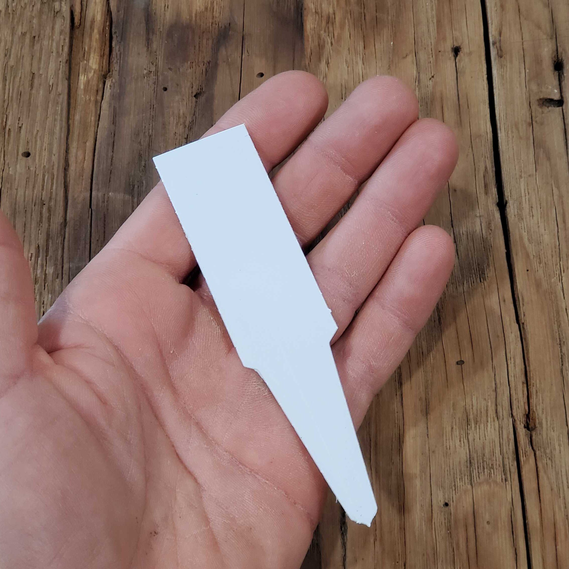 Plastic Plant Markers - Seeds for Generations A hand holds a white, flat Plastic Plant Marker Label 4.5" with a pointed end, positioned over a wooden surface.