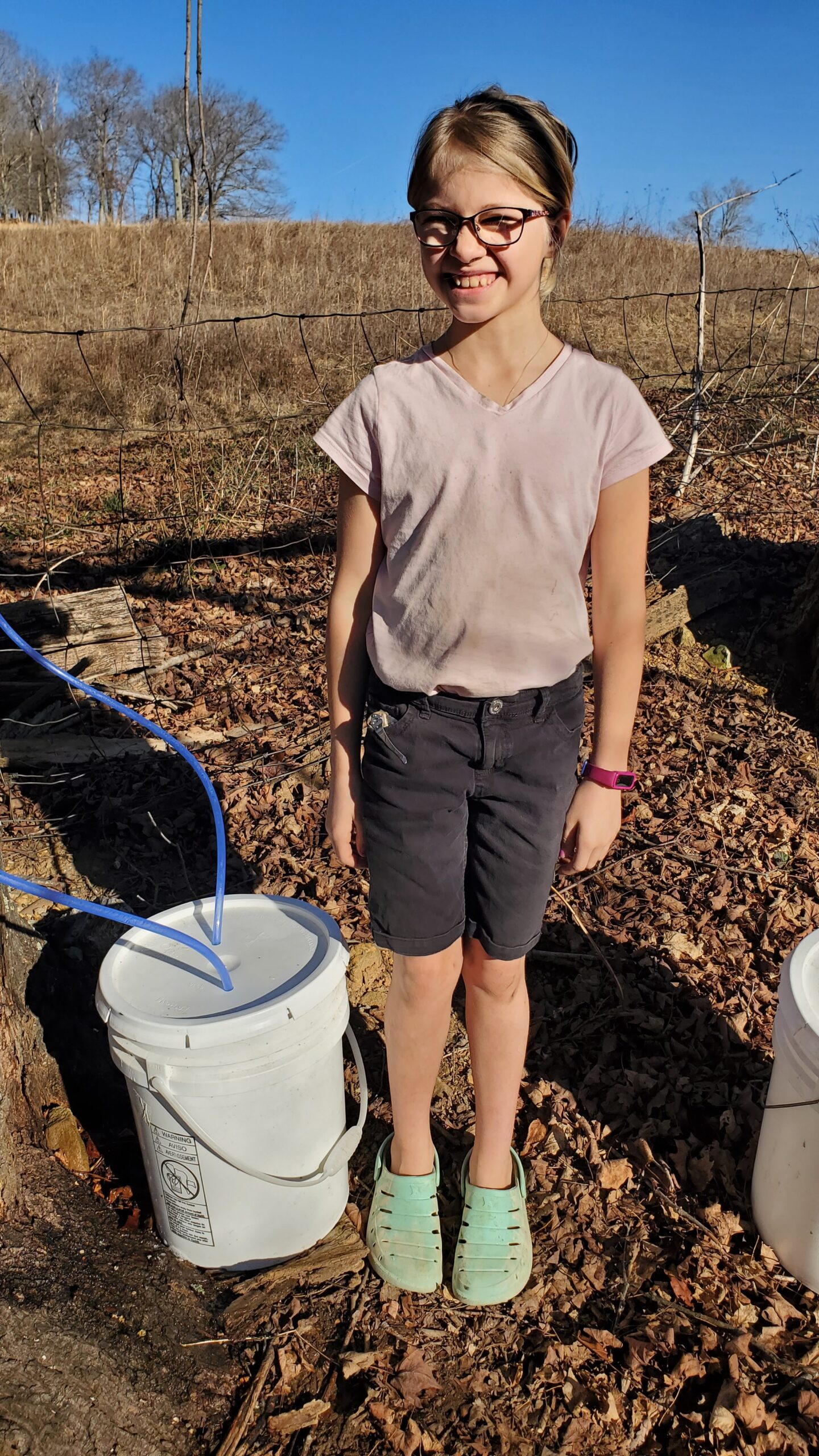 A girl standing next to two buckets in the woods.