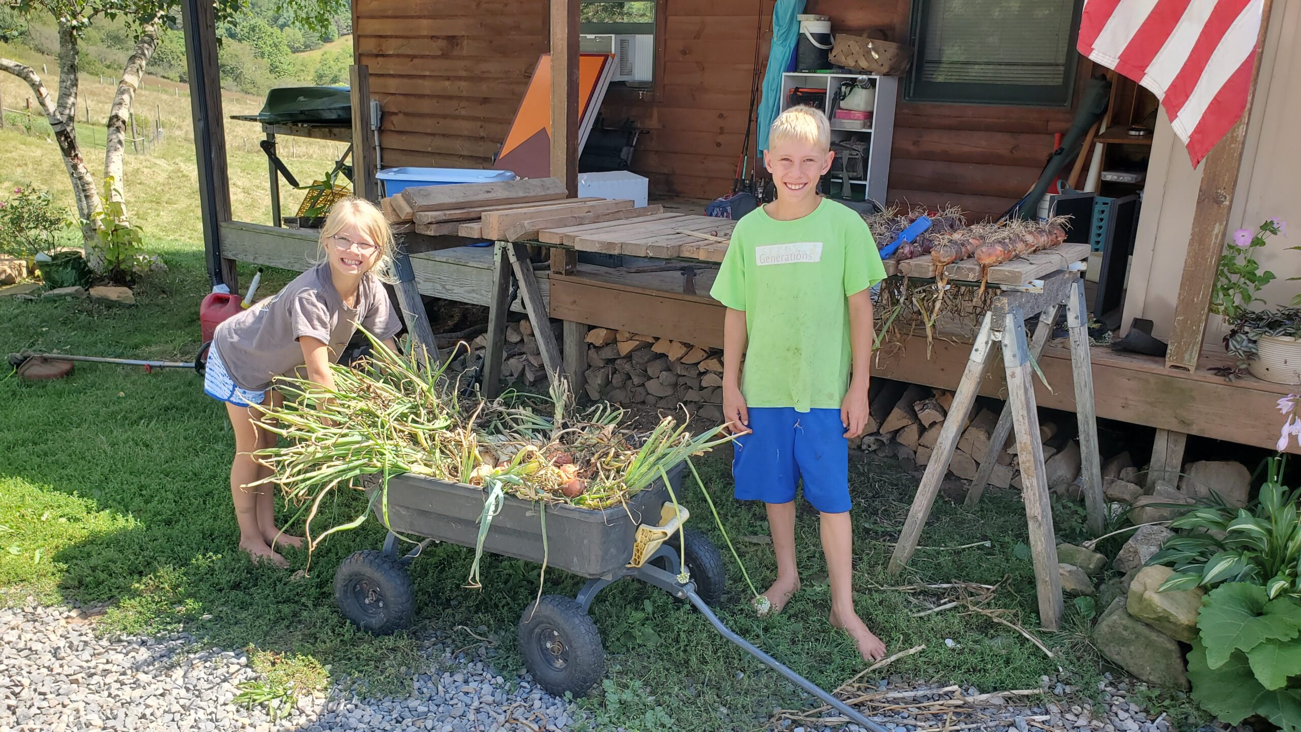Two children smiling and standing next to a cart full of freshly harvested onions in front of a cabin with an American flag.