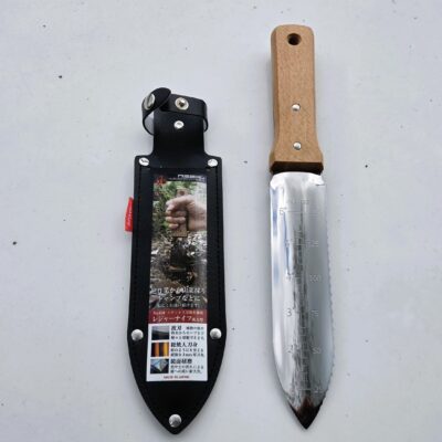 A Hori Hori Knife made of stainless steel with measurement markings rests beside its black leather sheath, which displays a product label in Japanese.