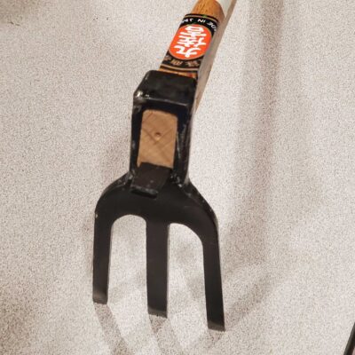 The Kuwa Hoe, featuring a wooden handle and metal prongs, is placed on a speckled gray surface.