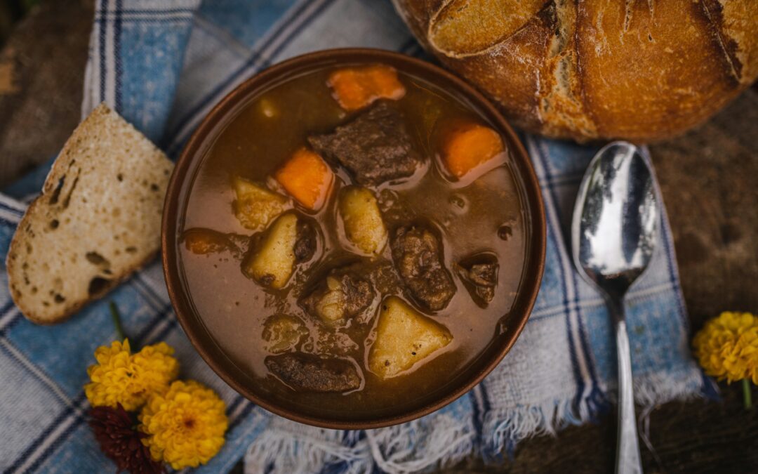 Bowl of beef stew with carrots and potatoes on a checkered cloth, surrounded by a spoon, bread, and flowers.