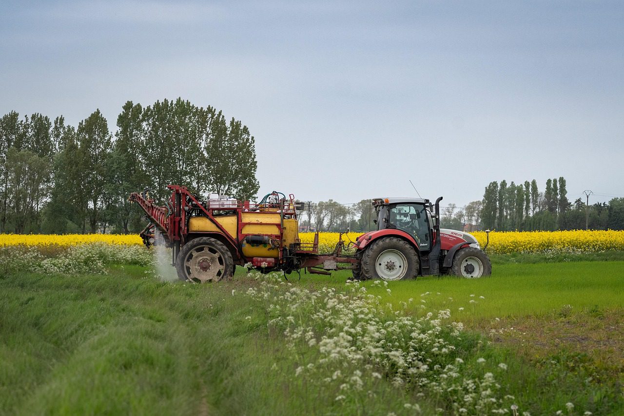 A red tractor with a yellow sprayer attachment moves through a green field, with trees and a blooming yellow flower field in the background.