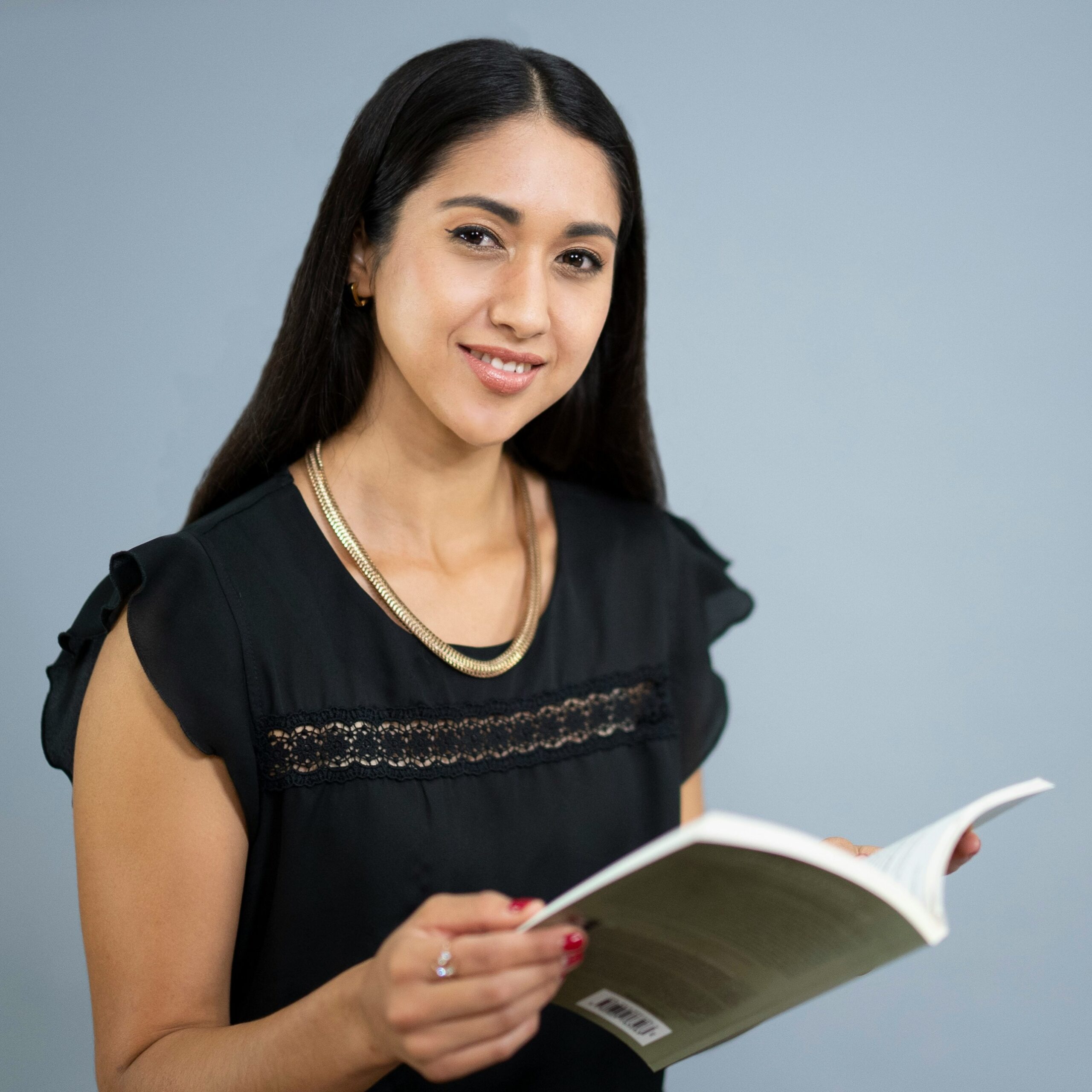 Woman with long dark hair wearing a black blouse and gold necklace holds an open book and smiles at the camera against a plain background.