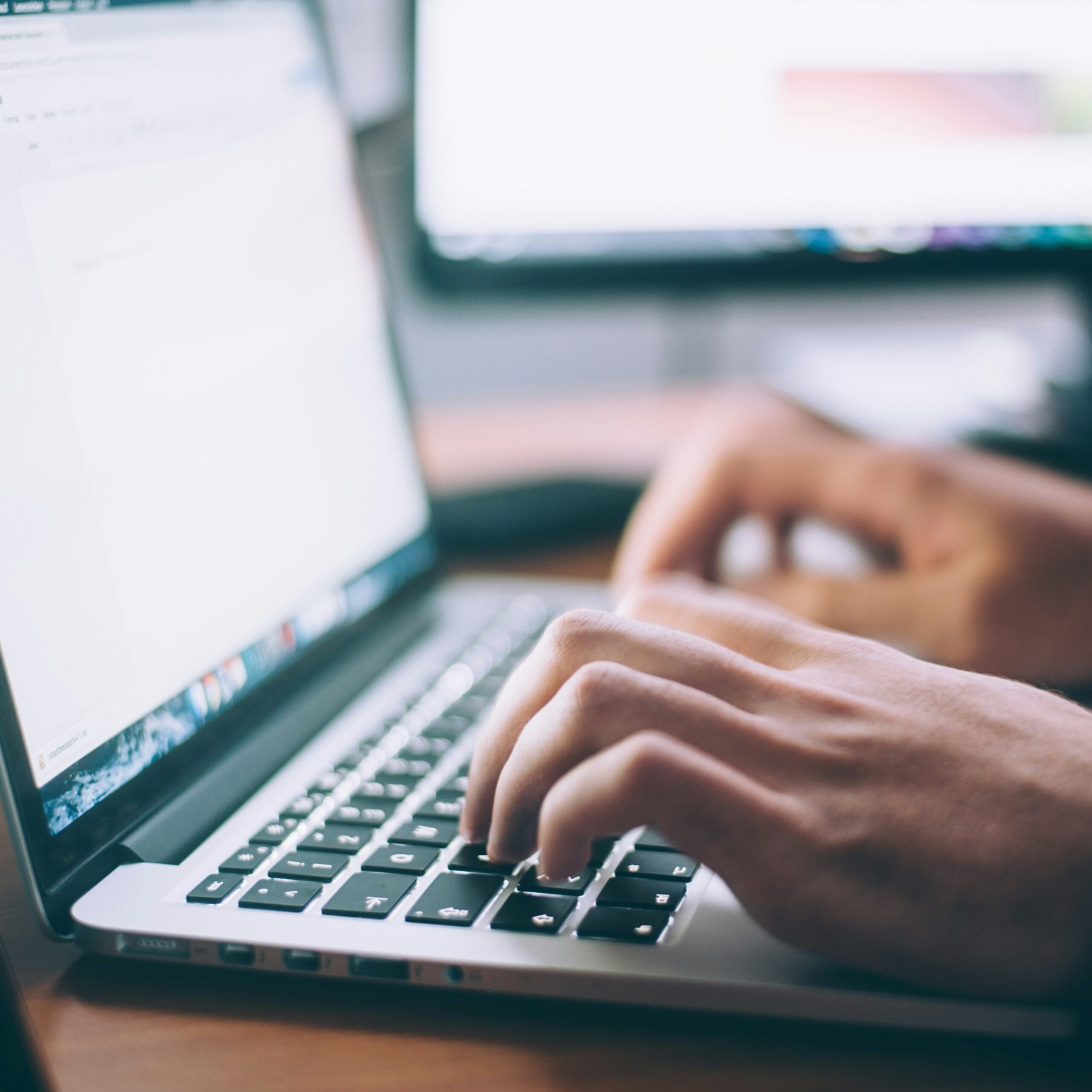 Close-up of a person's hands typing on a laptop keyboard, with a blurred computer monitor in the background.