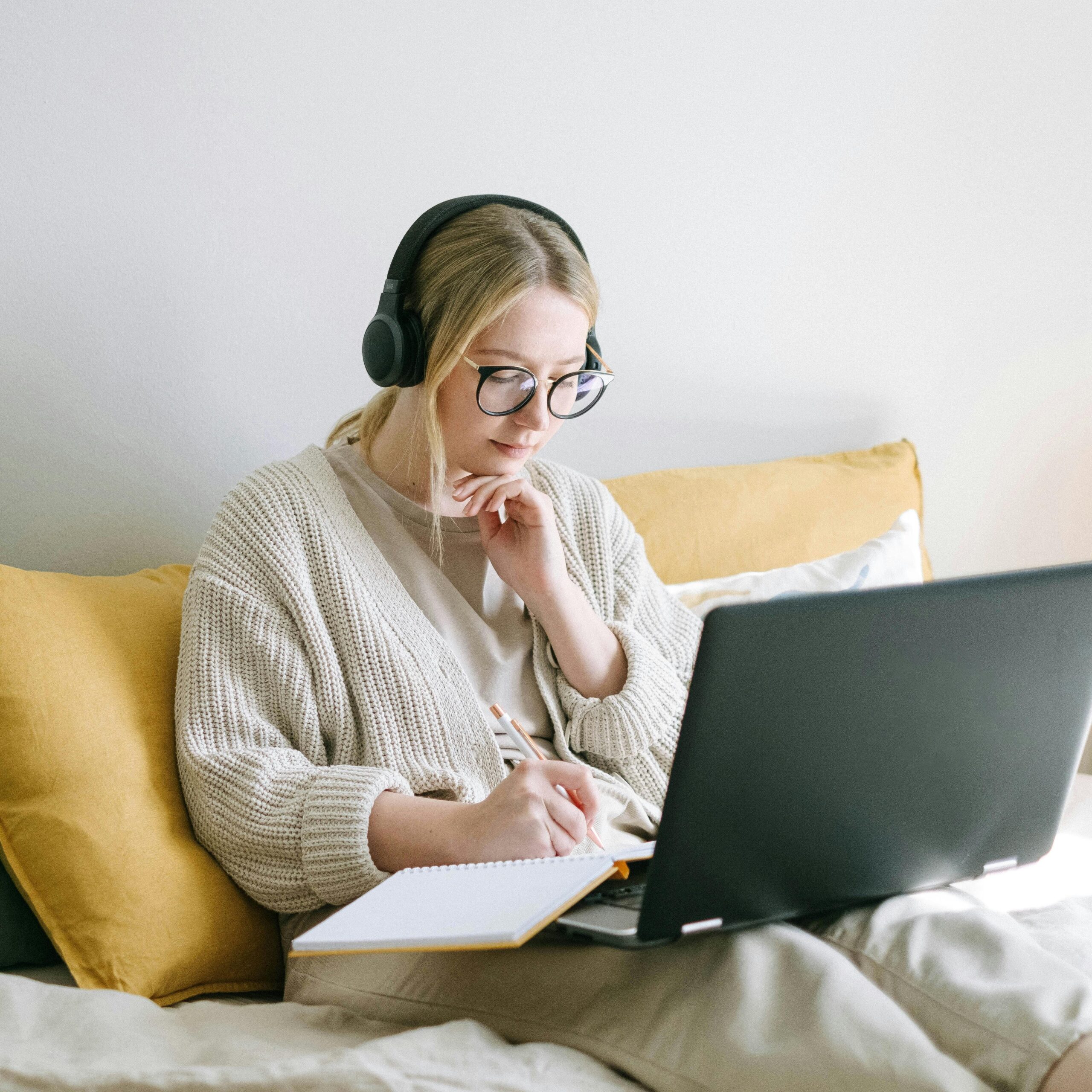 A person wearing headphones and glasses sits on a bed with yellow pillows, writing in a notebook while using a laptop.