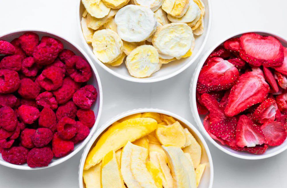 Four bowls filled with freeze-dried raspberries, bananas, strawberries, and mango slices arranged on a white surface.
