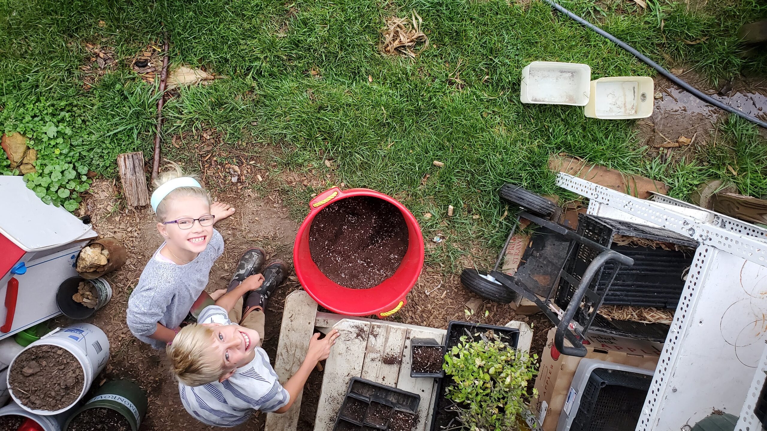 Two children smile up at the camera while working in a garden, surrounded by soil, containers, and gardening tools on a grassy area.
