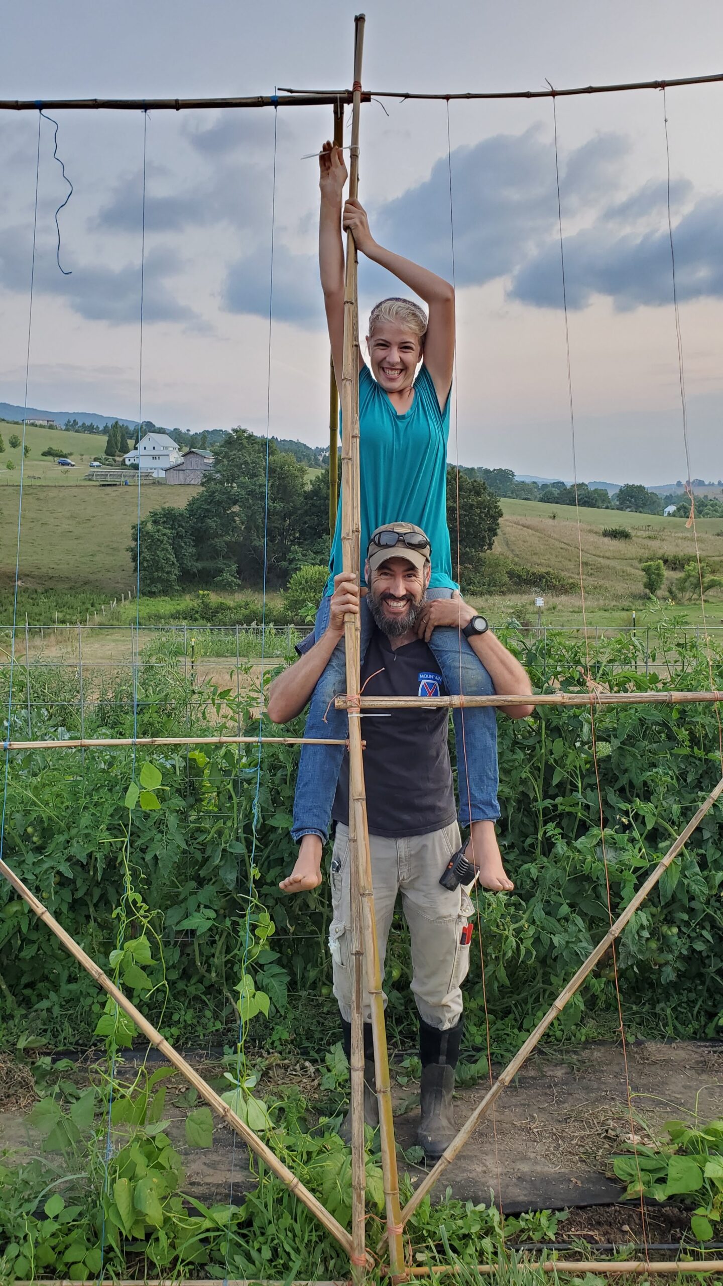 An adult stands in a garden, holding onto a bamboo trellis, while a child sits on their shoulders and holds onto the top of the structure. Fields and houses are visible in the background.