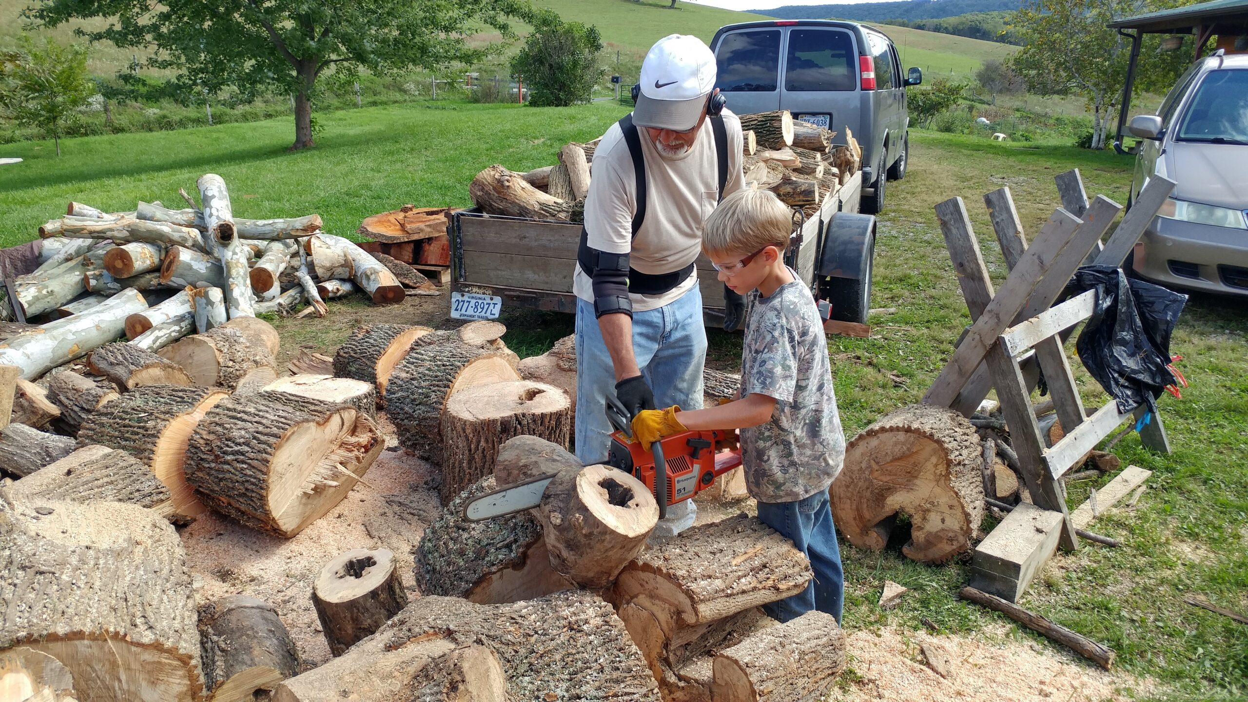 An older man and a boy cut large logs with a chainsaw outdoors, surrounded by chopped wood and vehicles in a grassy rural area.