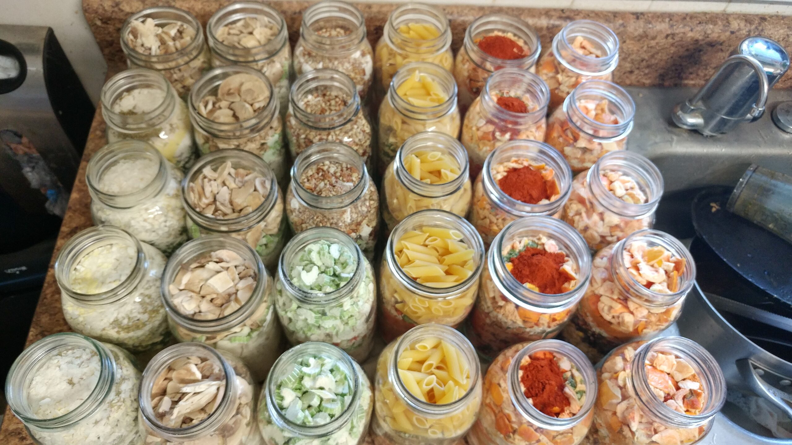 Several glass jars filled with various dried ingredients including vegetables, pasta, and seasonings, are arranged in rows on a kitchen countertop near a sink.