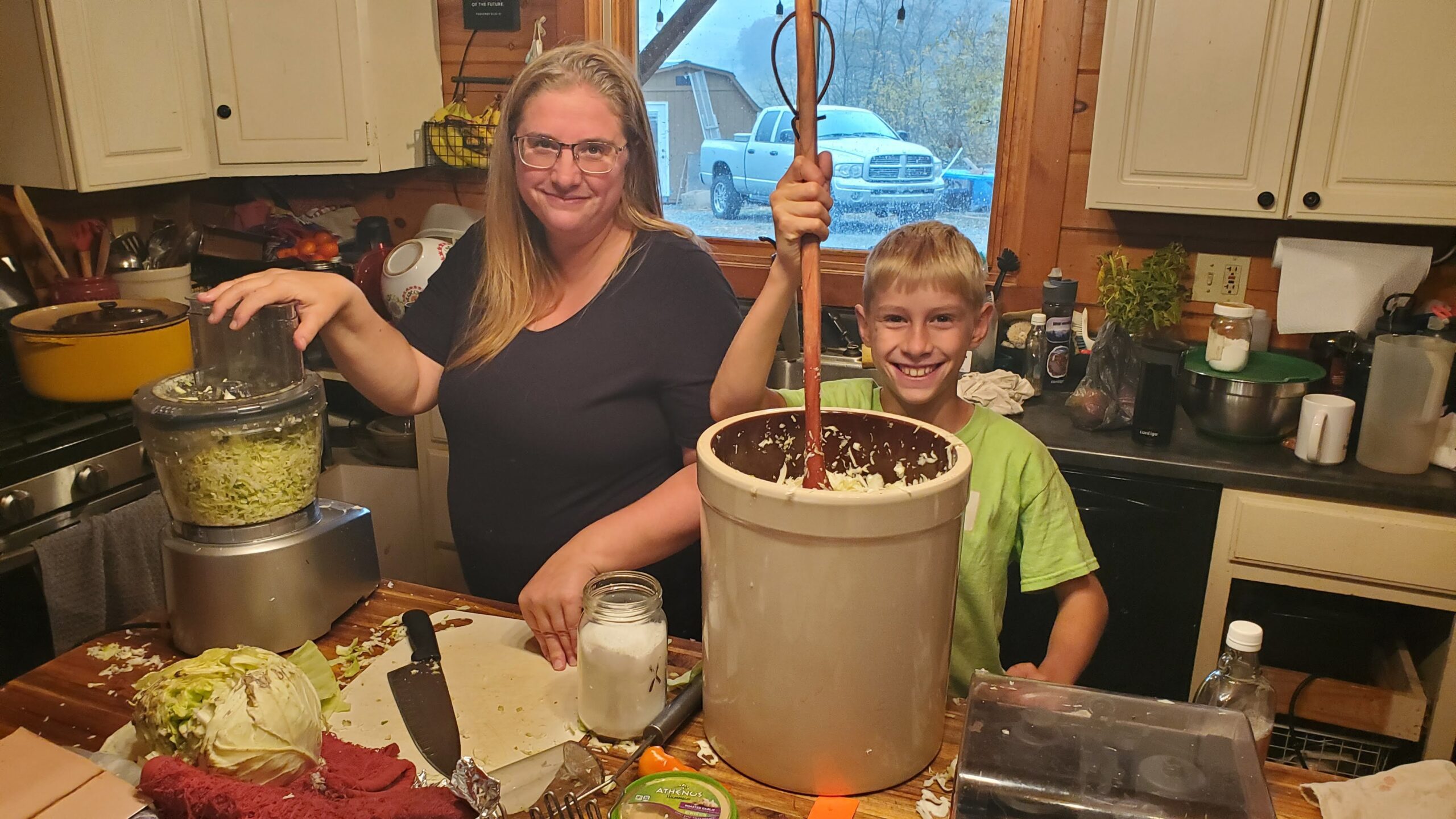 A woman uses a food processor while a smiling boy stirs shredded cabbage in a large bucket in a kitchen with various utensils and ingredients on the counter.