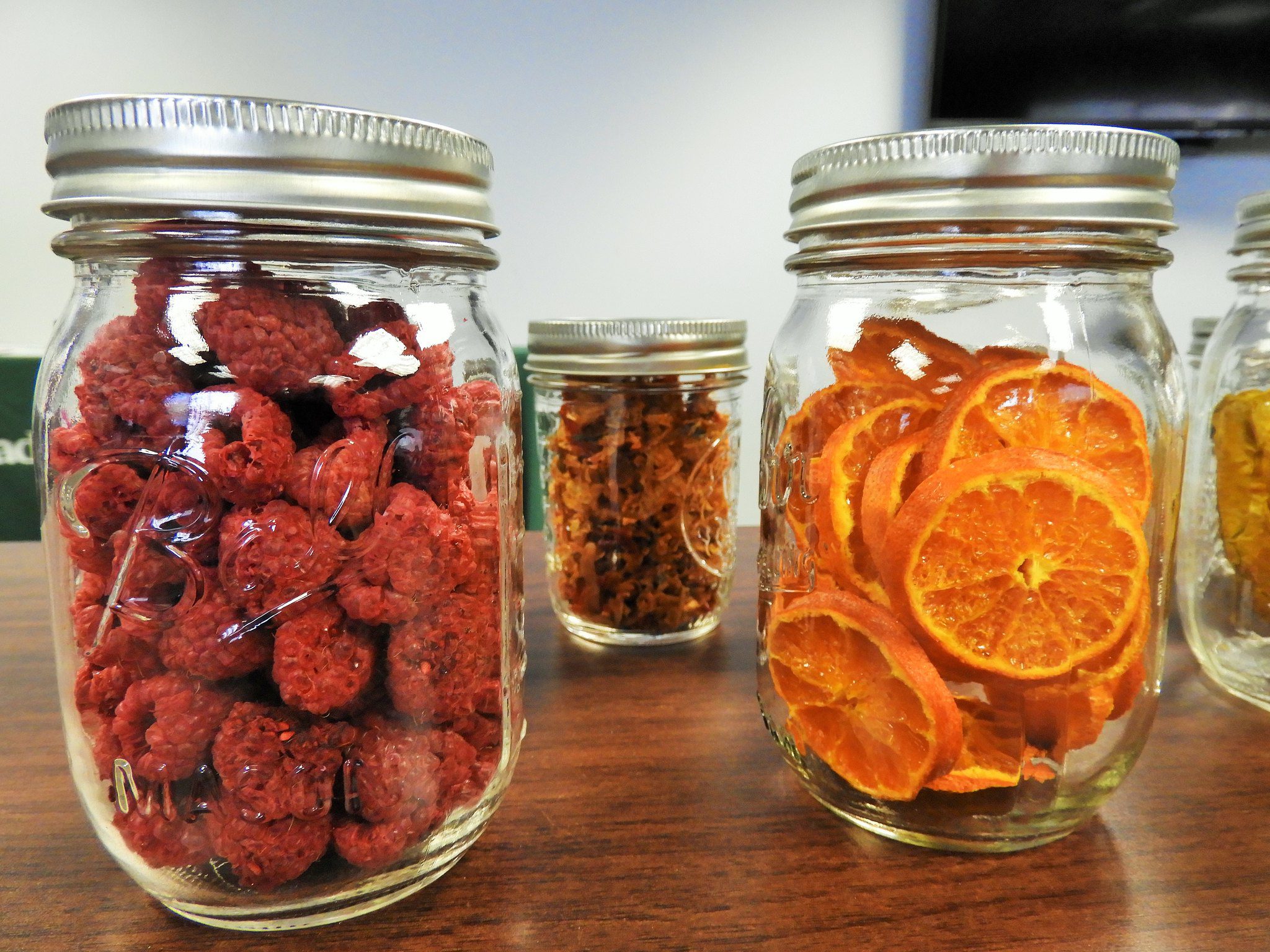 Three glass jars containing dried raspberries, orange slices, and an unidentified dried food item, displayed on a wooden surface.